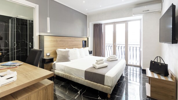 A modern hotel room with a large bed, a wooden desk, a TV, and a balcony with a view. - Hotel Bella Napoli (Naples)
