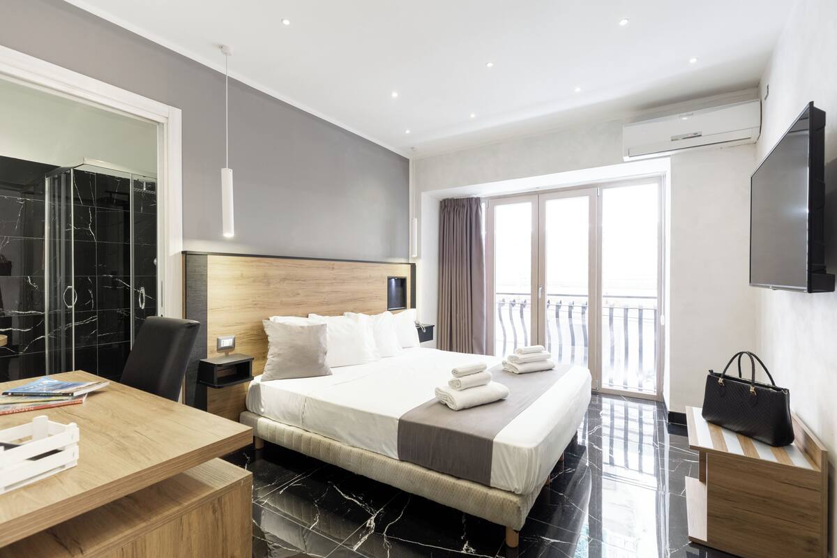 A modern hotel room with a large bed, a wooden desk, a TV, and a balcony with a view.