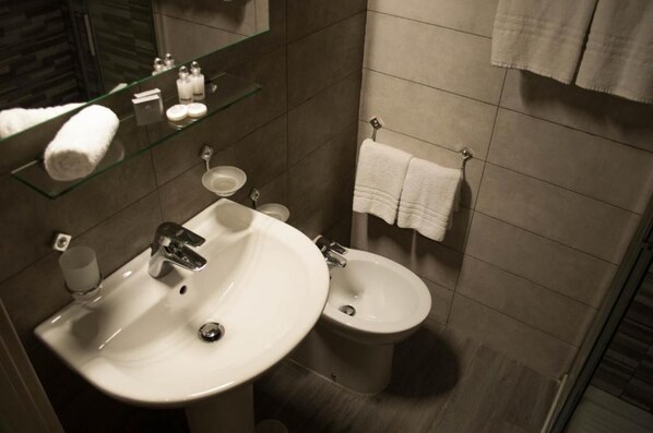 Une salle de bain moderne avec un lavabo blanc, un bidet et une étagère en verre où sont rangés les articles de toilette.