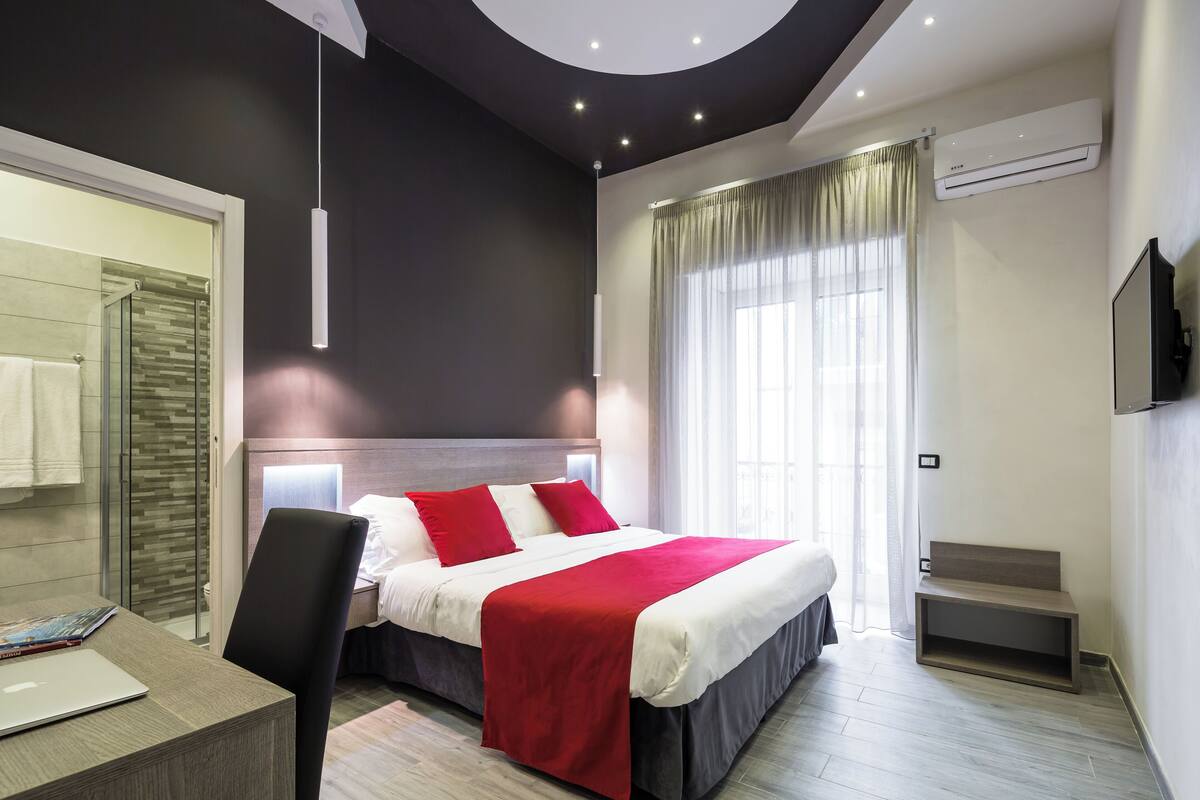 A modern hotel room with a bed, a desk, a chair, a shower area, and a large window with curtains.