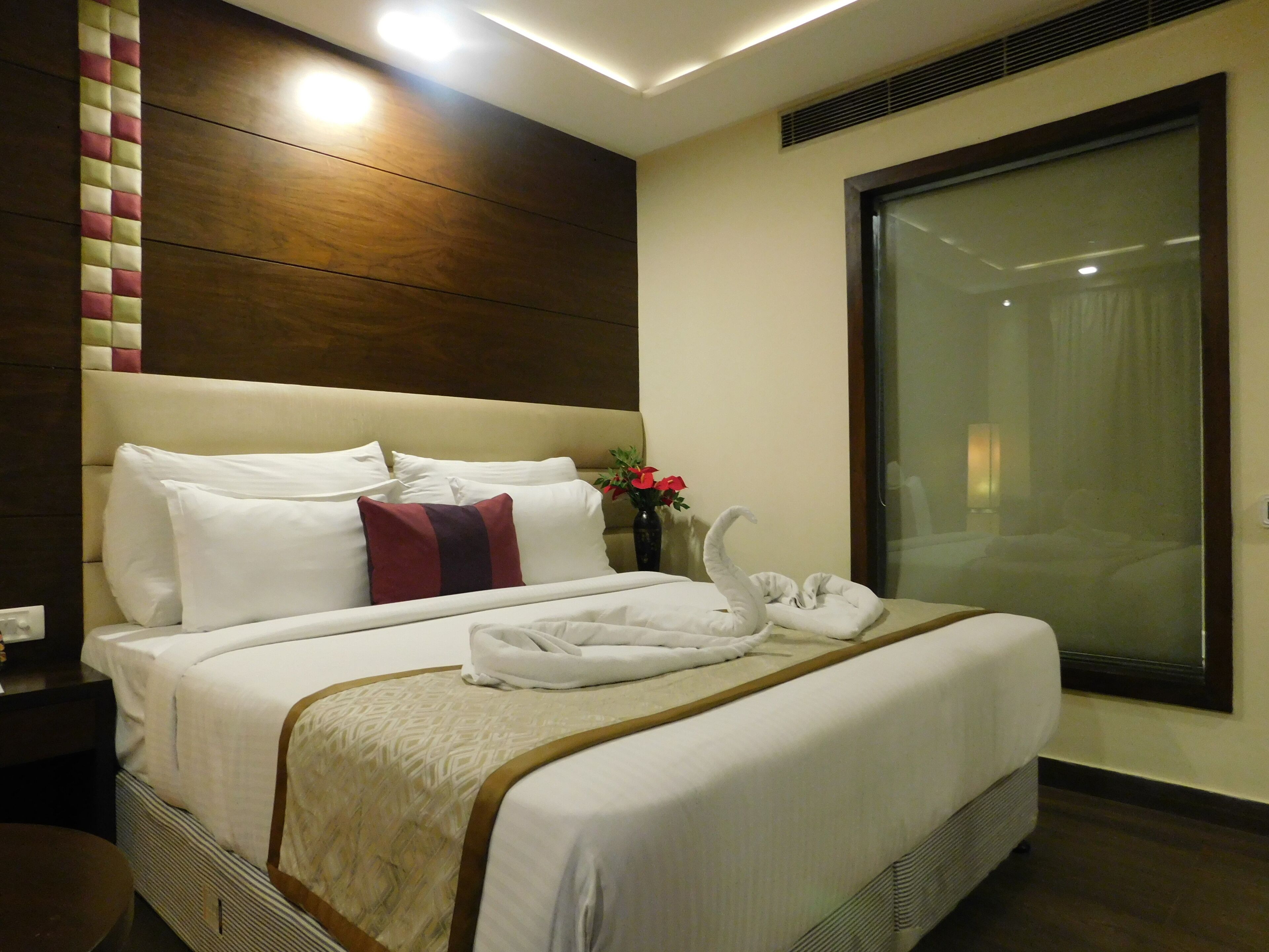 Ein Hotelzimmer mit einem großen Bett, einem hölzernen Kopfteil, einer Vase mit Blumen und einem Fenster mit Vorhängen.
