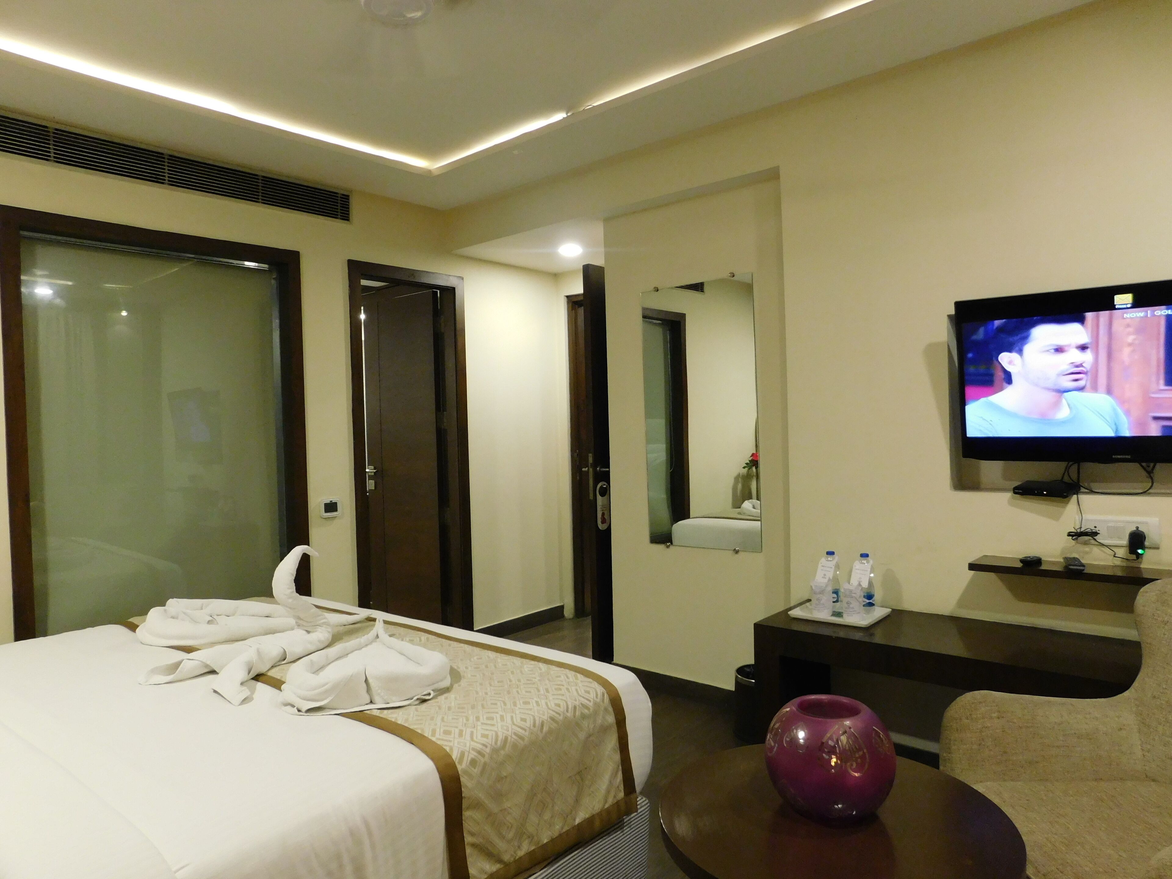 A hotel room with a bed, a television, a mirror, a small table with a vase, and a chair.