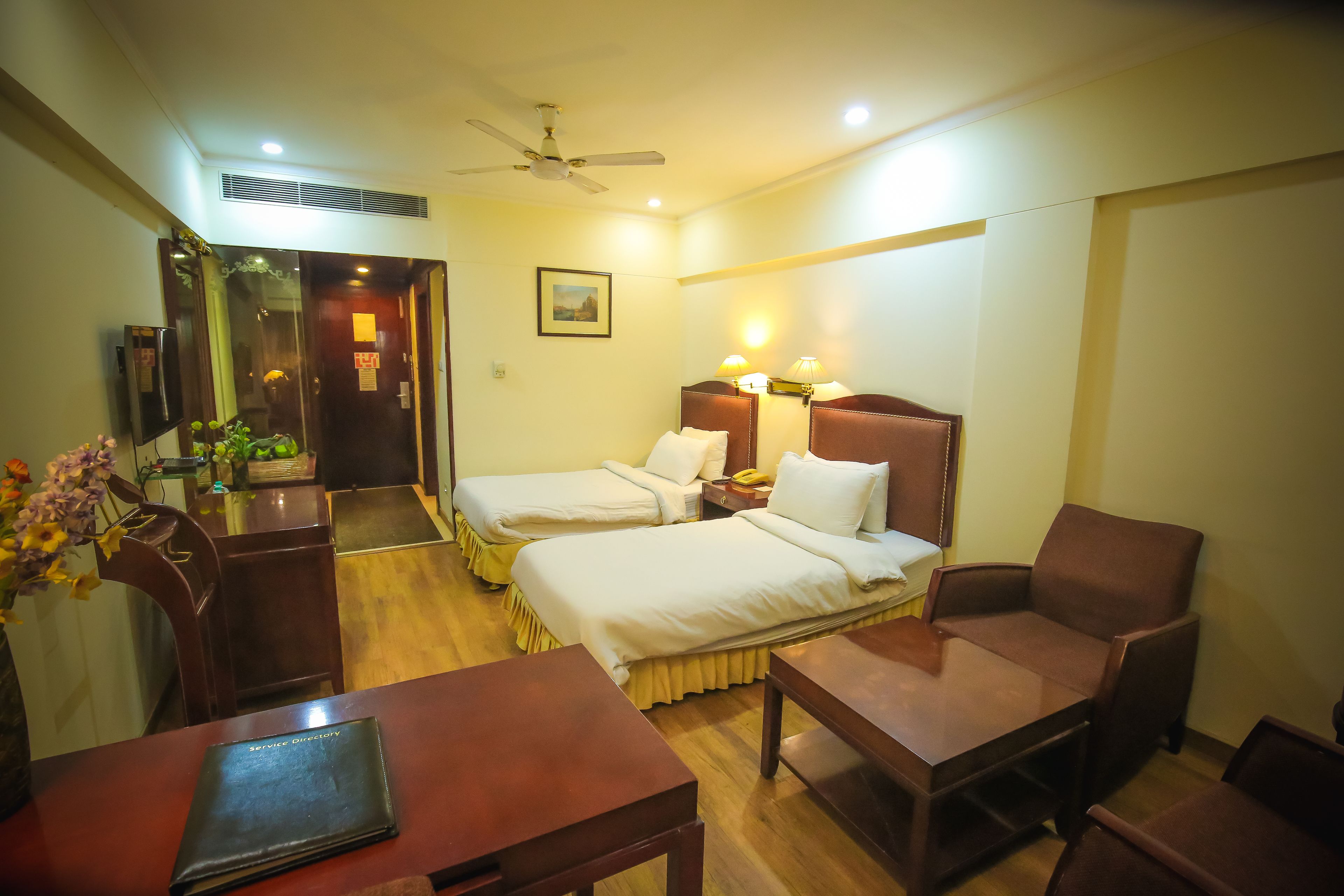A hotel room with two beds, a desk, a chair, a table, a ceiling fan, and a wall-mounted TV.