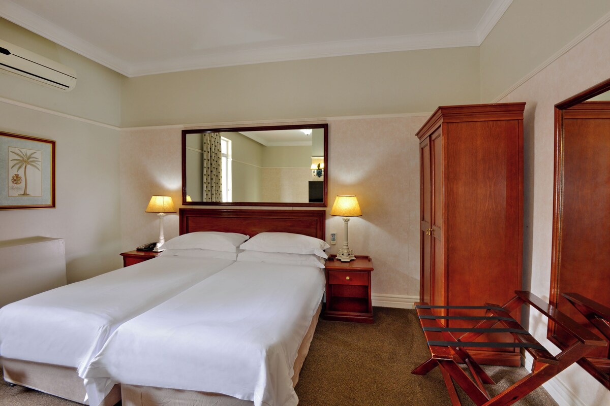 A hotel room with two beds, a wooden wardrobe, a desk with a lamp, and a mirror on the wall.