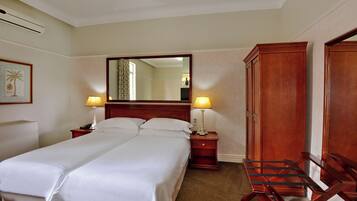A hotel room with two beds, a wooden wardrobe, a desk with a lamp, and a mirror on the wall.