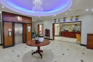 A hotel lobby with a reception desk, a chandelier, a wooden reception desk, and a table with a vase. - Balmoral (Durban)
