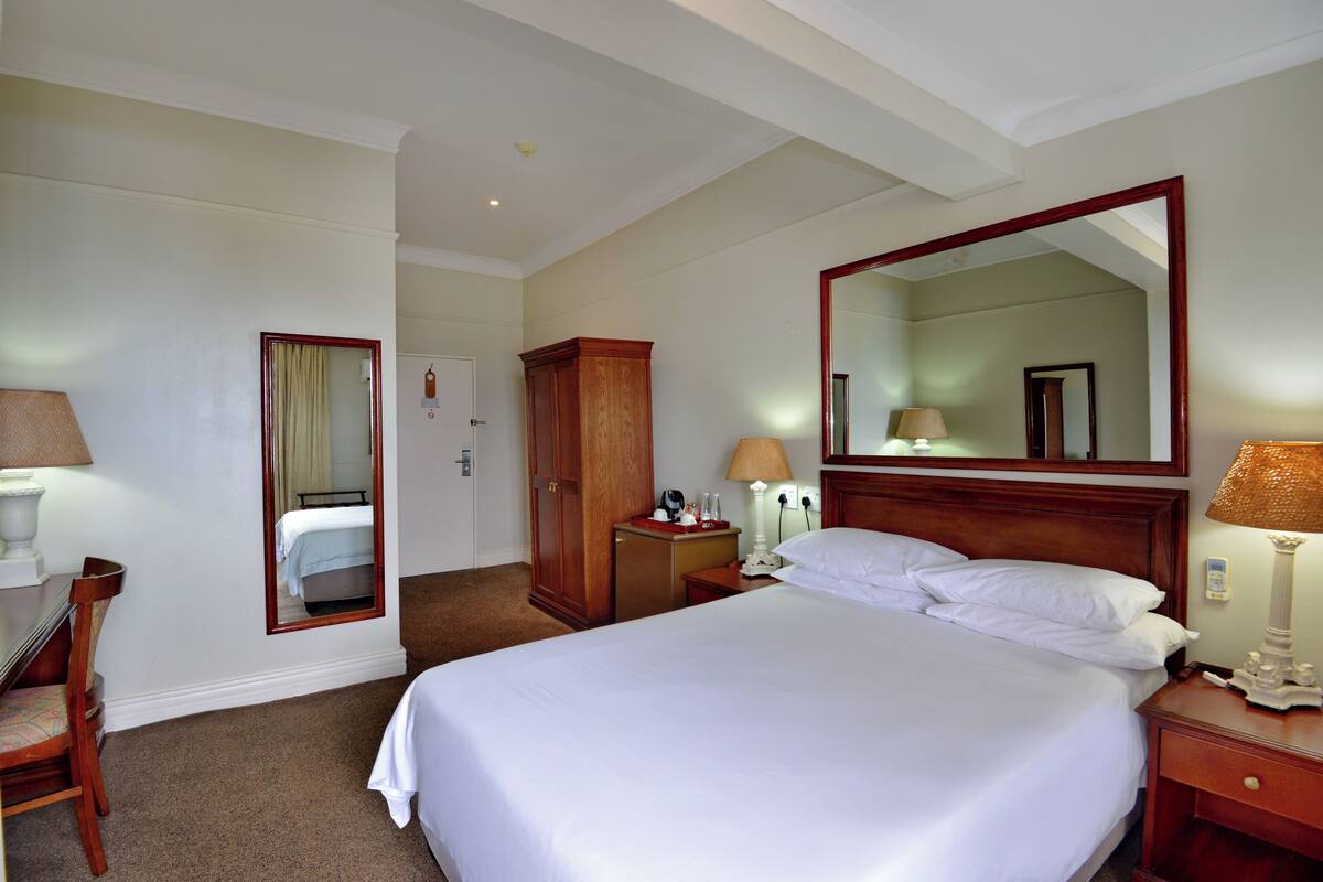 Una habitación de hotel con una cama grande, un armario de madera, un escritorio con una silla, una mesita de noche con una lámpara y un espejo en la pared.
