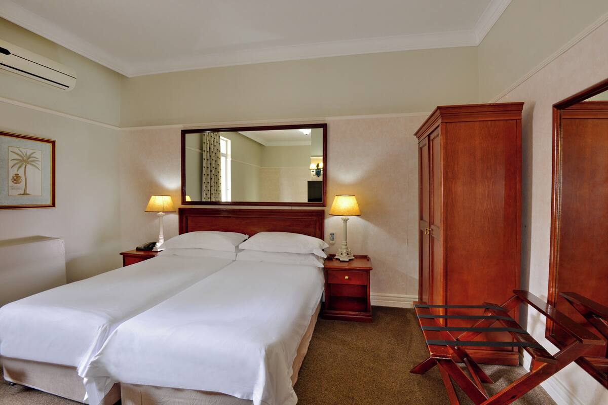 Una habitación de hotel con una cama doble, dos mesitas de noche con lámparas, un espejo grande, un armario de madera y un cuadro enmarcado en la pared.