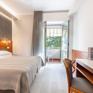 A hotel room with a bed, a desk, and a chair. There is a balcony with a view of greenery.