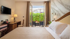 A hotel room with a bed, a desk, a TV, and a view of an outdoor area with a table and chairs. - Nicoletta Hotel (Baunei)