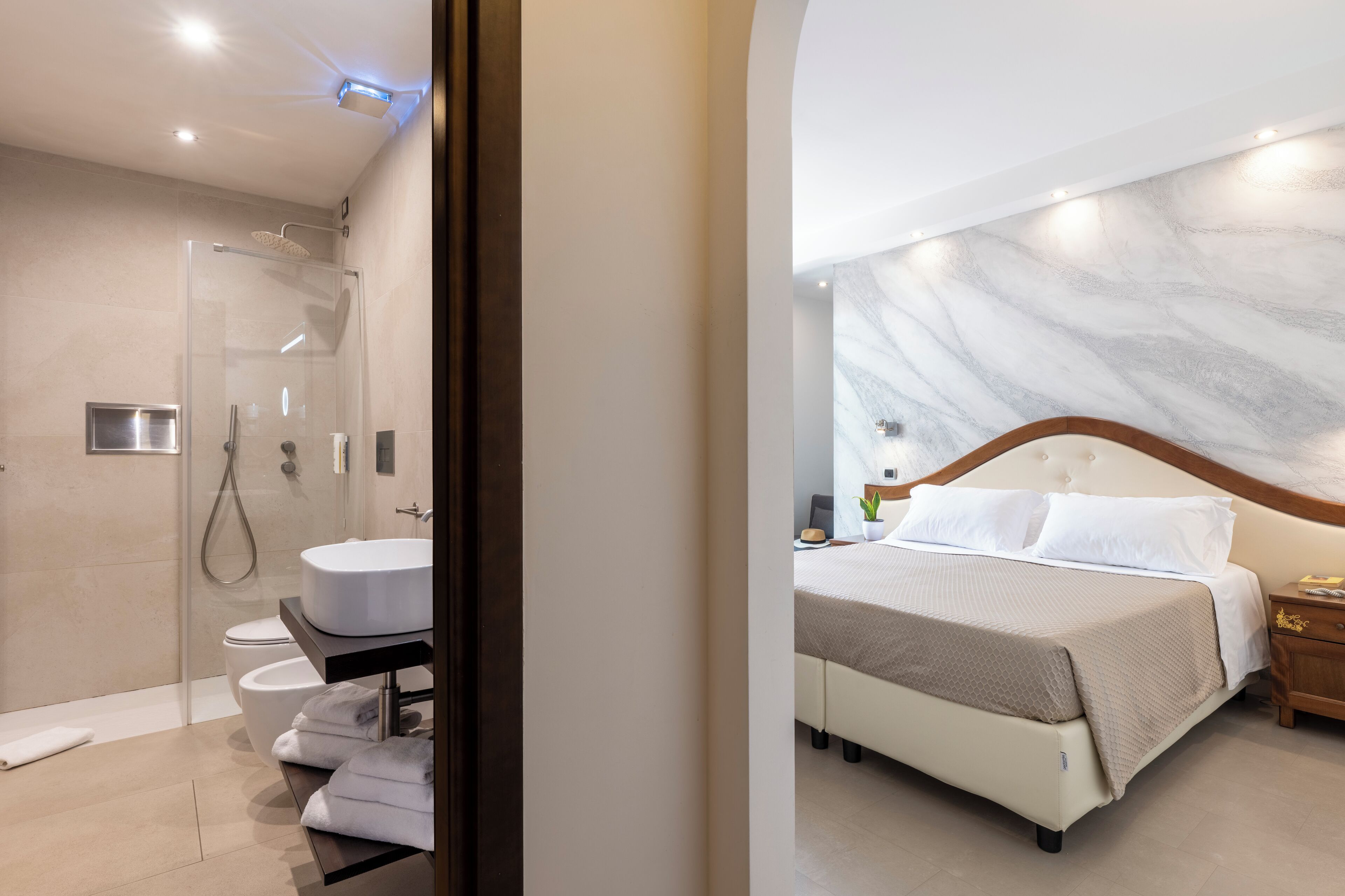 A modern hotel room with a glass-enclosed shower, a white sink, a bed with a wooden headboard, and a marble-patterned wall.