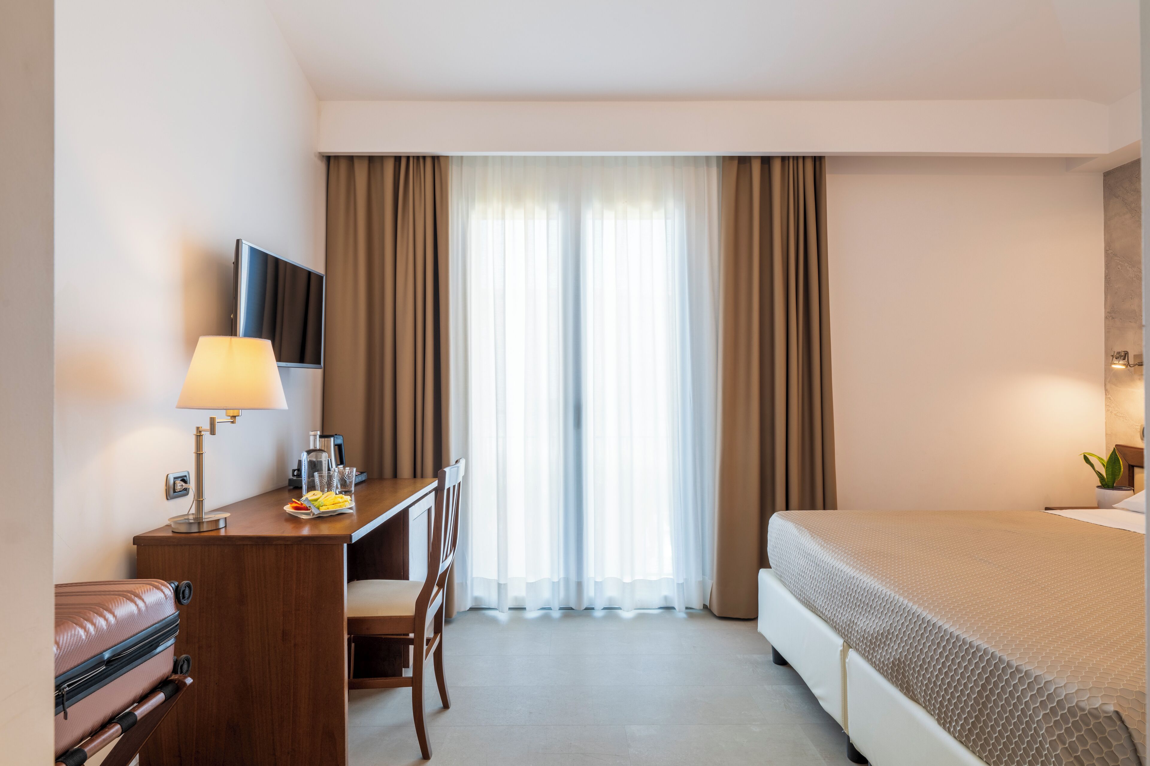 A hotel room with a bed, a desk with a lamp, a chair, a TV, and a window with curtains.