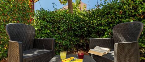 Une terrasse avec des chaises en osier et une table ronde, une assiette de fruits et deux boissons.