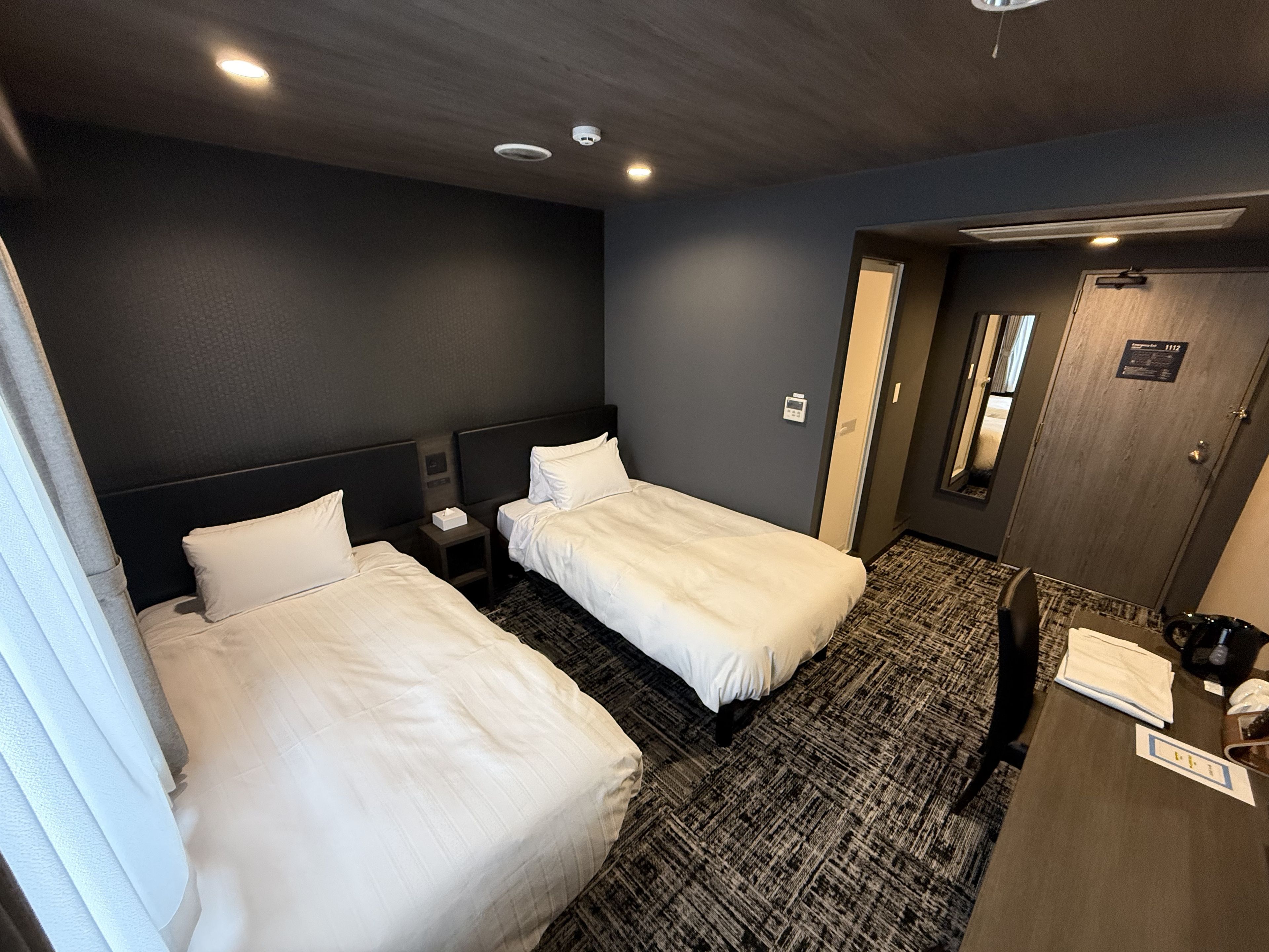 Habitación de hotel con dos camas, un escritorio, una silla, una puerta y una mesita con un teléfono y papeles.