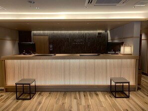 Un área de recepción moderna de hotel con un mostrador de madera, dos taburetes de bar, un televisor de pantalla plana y un panel de control montado en la pared.