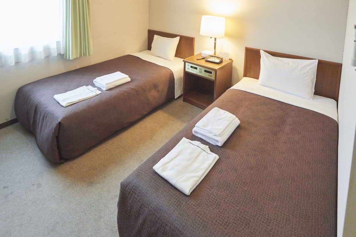 A hotel room with two beds, each with a brown bedspread and white pillows. There is a nightstand with a lamp and a phone between the beds.