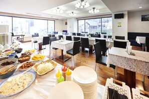 A dining area with multiple tables and chairs, buffet with various food items, and large windows providing natural light.
