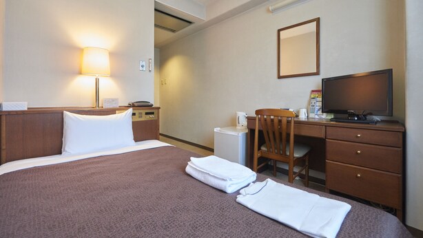 Quarto de hotel com cama, escrivaninha, cadeira, televisão e um pequeno frigorífico.