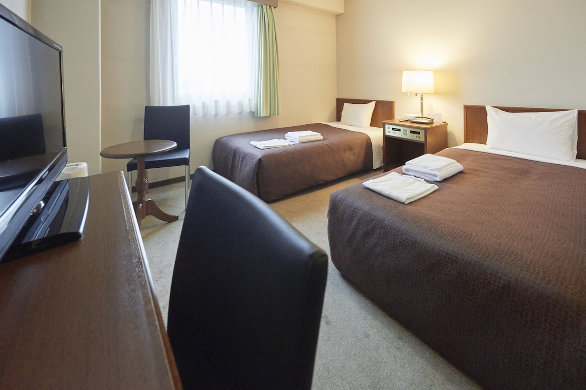 A hotel room with two beds, a desk with a TV, a chair, a bedside table with a lamp, and a window with curtains.