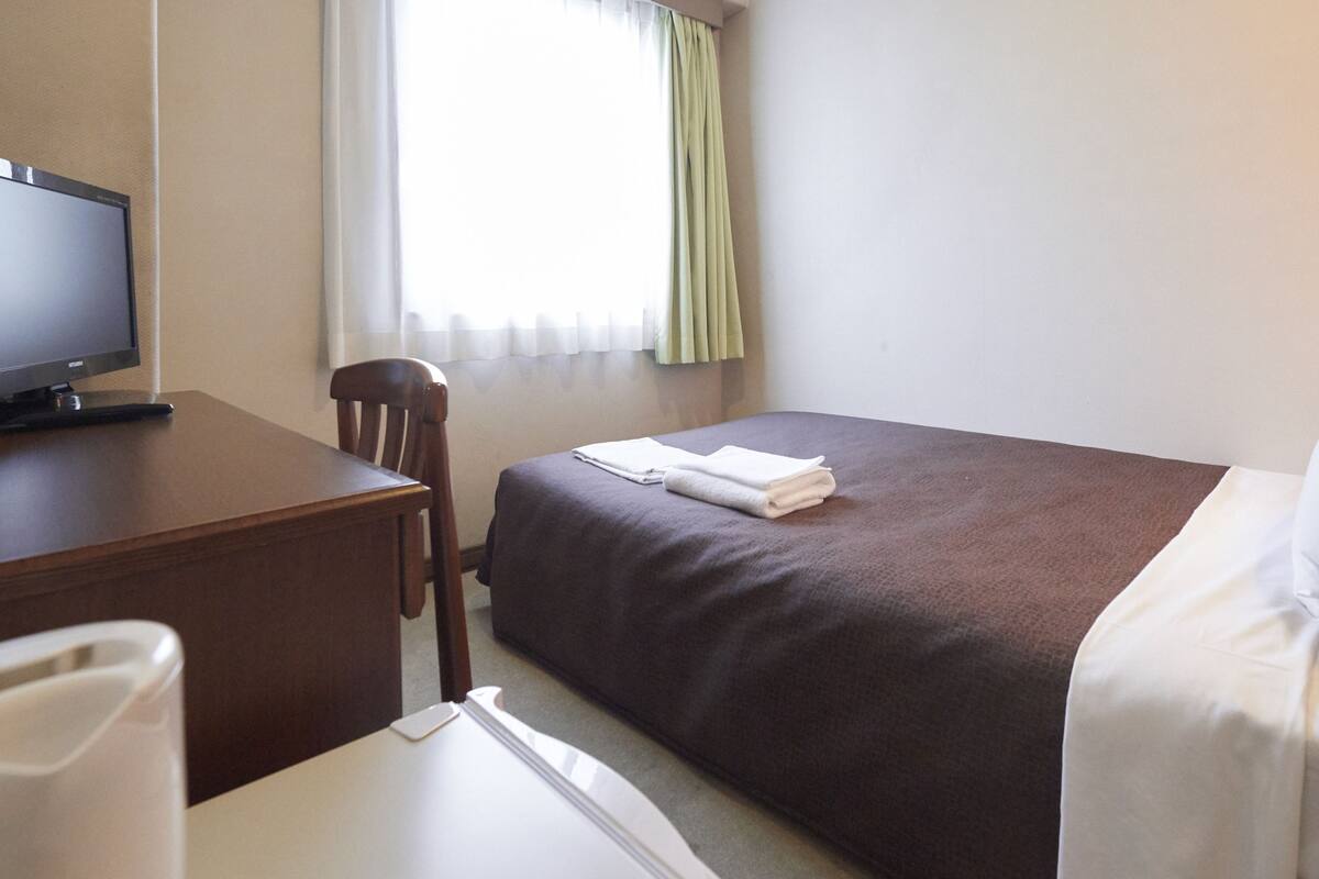 A hotel room with a bed, a desk with a television, a chair, and a lamp.