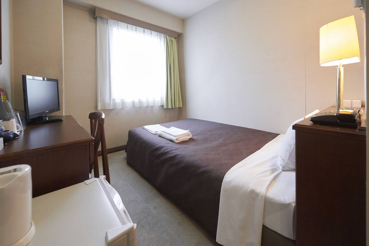 A hotel room with a bed, a desk with a computer, a lamp, and a window with curtains.