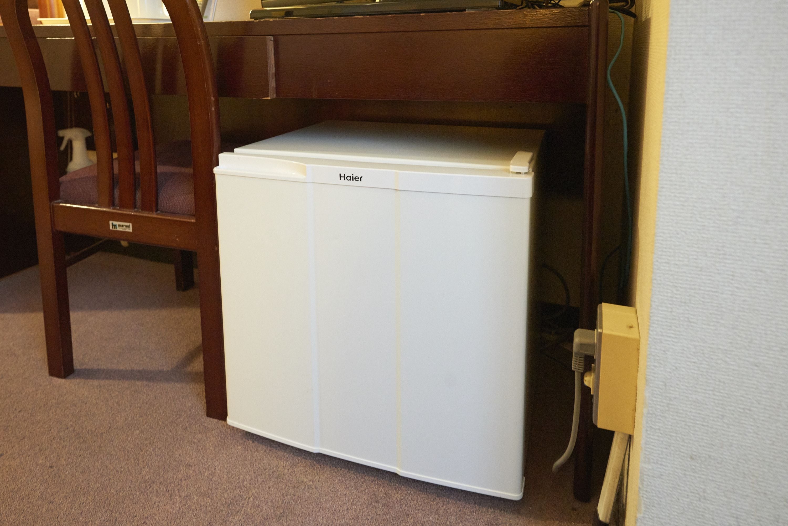 A compact white Haier refrigerator under a wooden desk with a chair, a power outlet, and a cable visible.