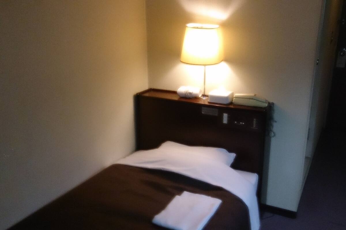 A hotel room with a single bed, a nightstand with a lamp, a telephone, and a small table with a towel and a bottle.