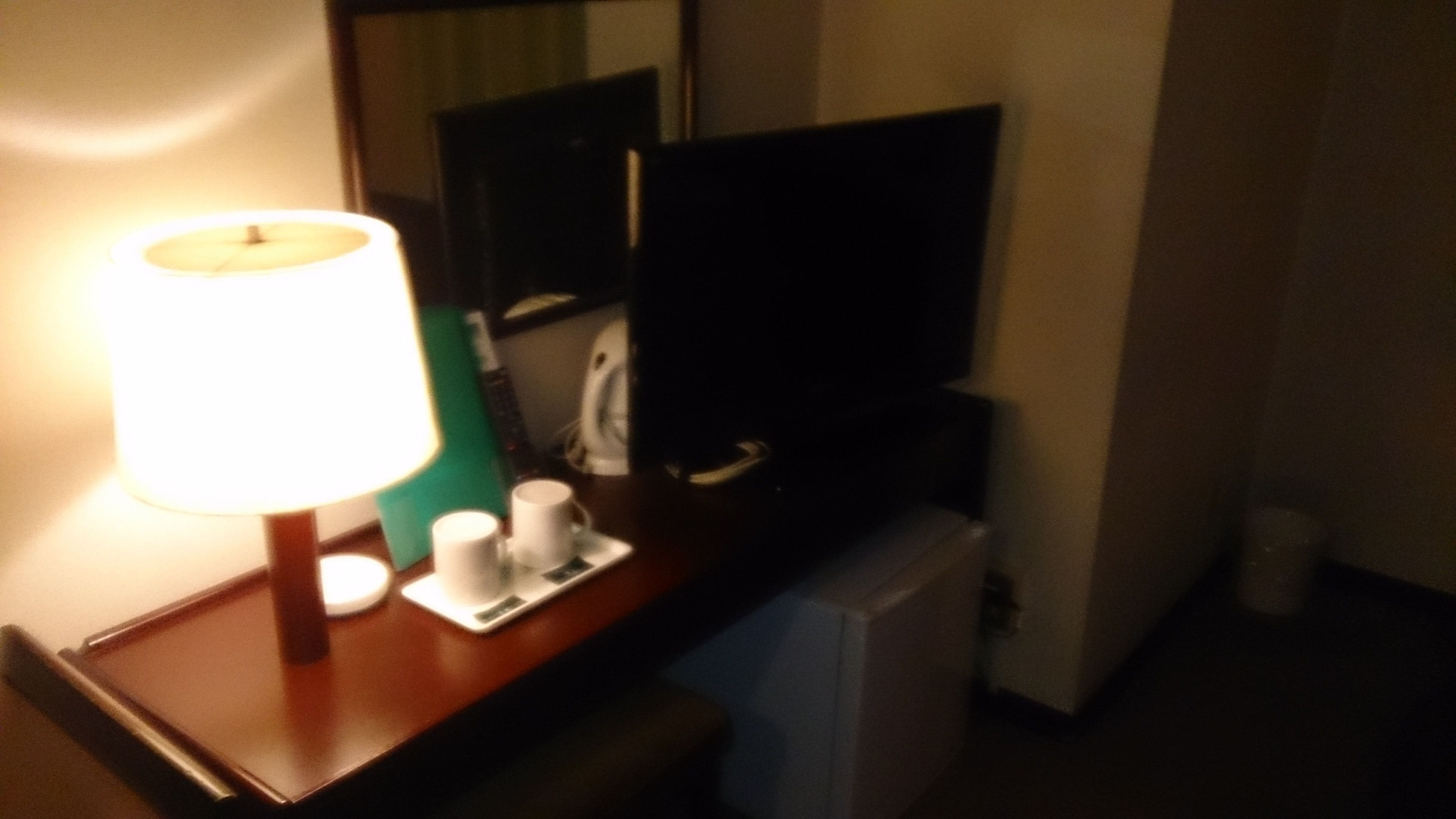 A hotel room with a desk, a lamp, a small refrigerator, and a mirror.