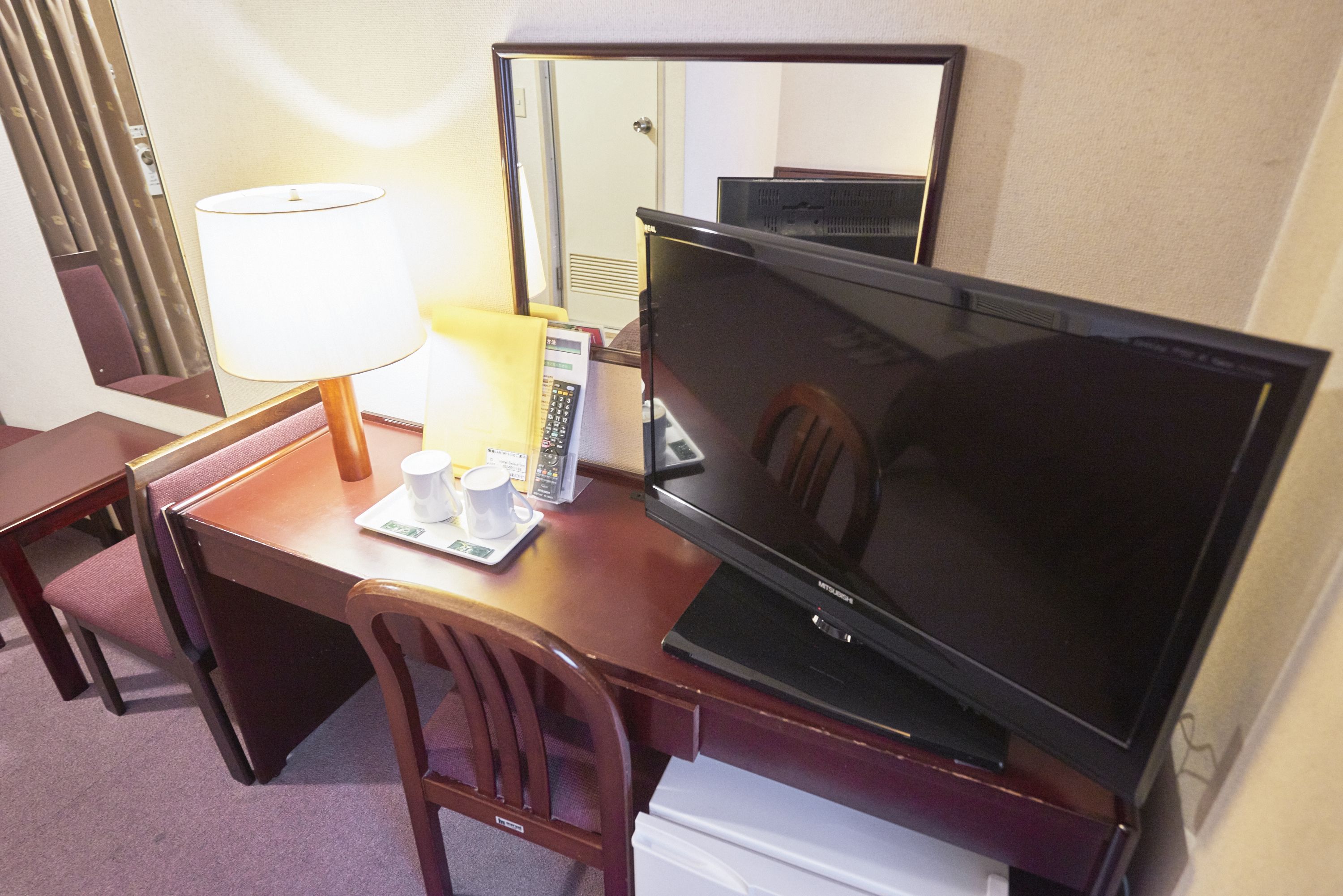 A hotel room with a desk, a television, a lamp, a telephone, and a mirror.