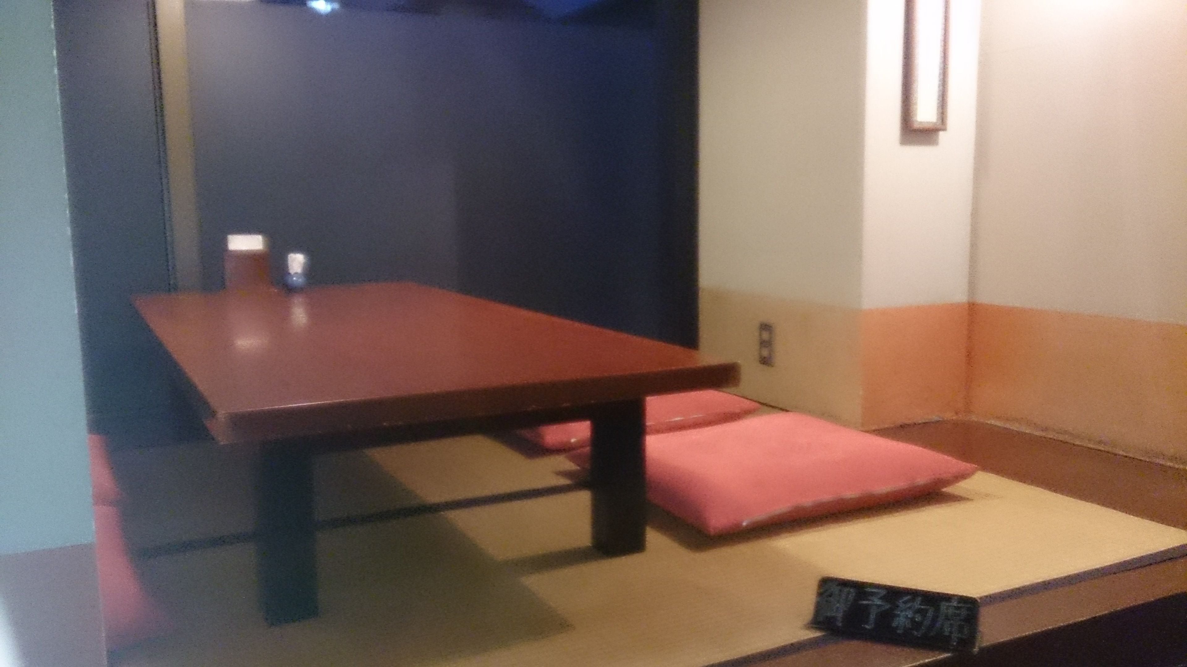 Una habitación con una mesa de madera, un cojín rosa, esteras de tatami y un pequeño estante con un cartel.