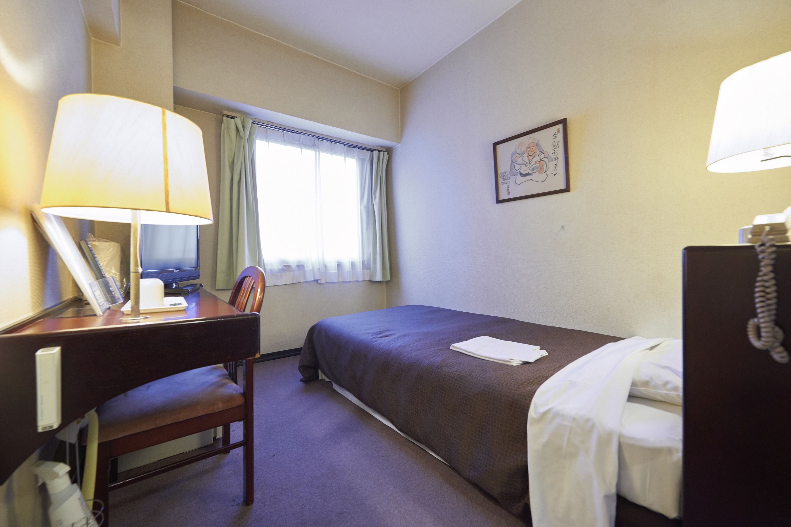 Hotelkamer met een bed, bureau met lamp, stoel, raam met gordijnen en een ingelijst kunstwerk aan de muur.