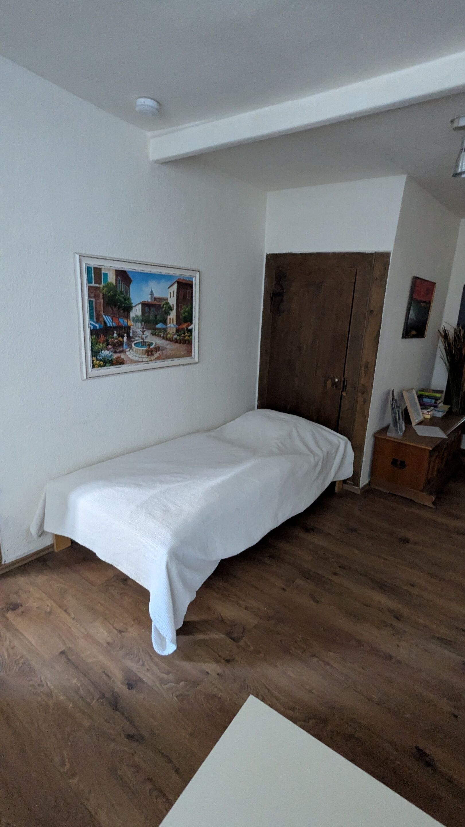 A bedroom with a single bed, a wooden wardrobe, a painting on the wall, and a small wooden cabinet.