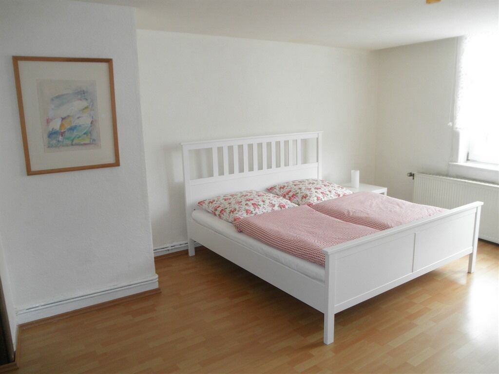 A white bed with floral pillows and a pink blanket in a room with wooden flooring and white walls.