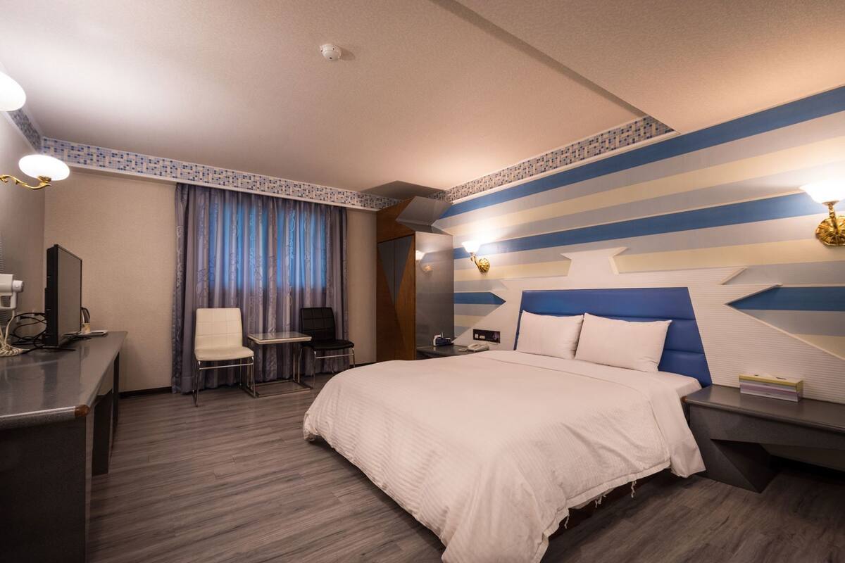A hotel room with a bed, desk, chair, and a desk lamp.