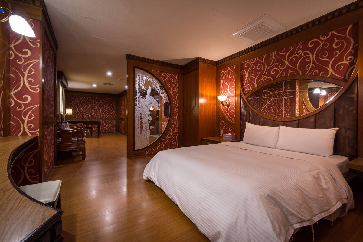 A hotel room with a large bed, wooden flooring, a mirror with a decorative frame, and a small table with a chair.
