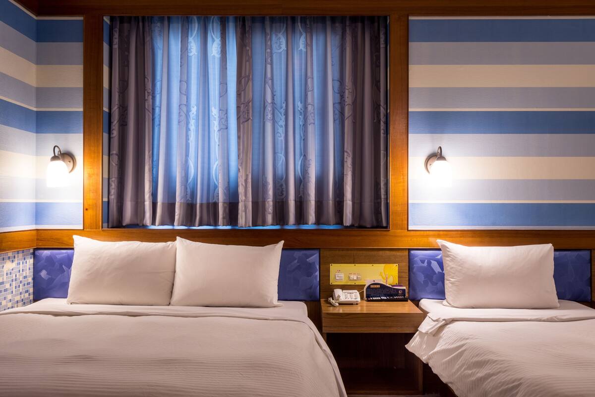A hotel room with two beds, a wooden headboard, a nightstand with a phone and a lamp, and a window with curtains.