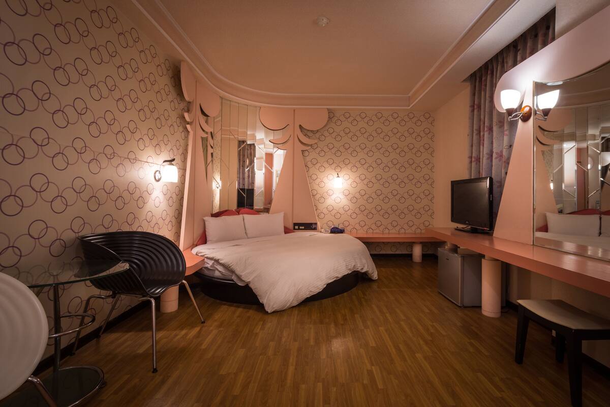 A modern hotel room with a wooden floor, a bed with a white duvet, a desk with a television, and a chair.