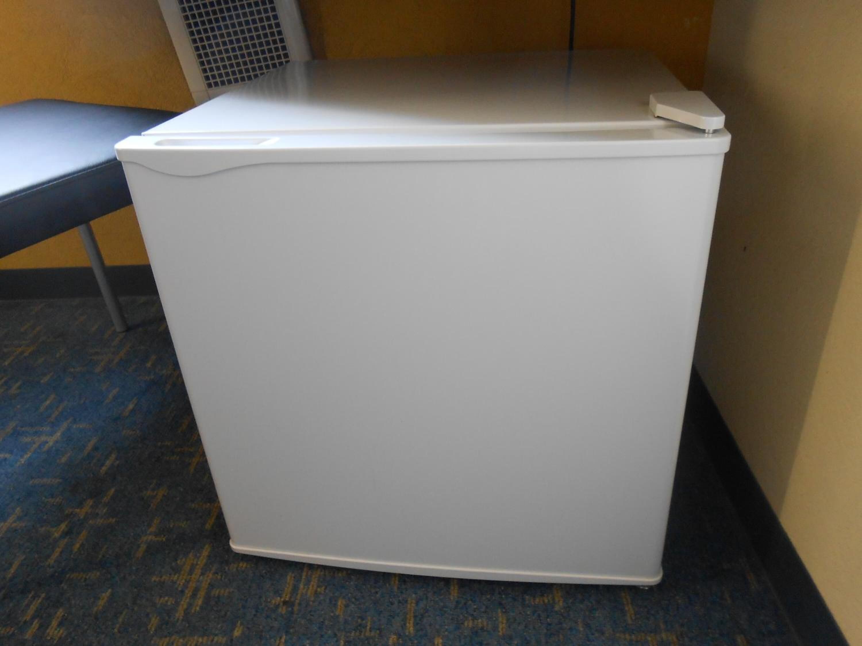 A compact, white, wall-mounted unit with a control panel on top, likely a laundry or utility appliance, in a room with a patterned carpet and a metal chair.