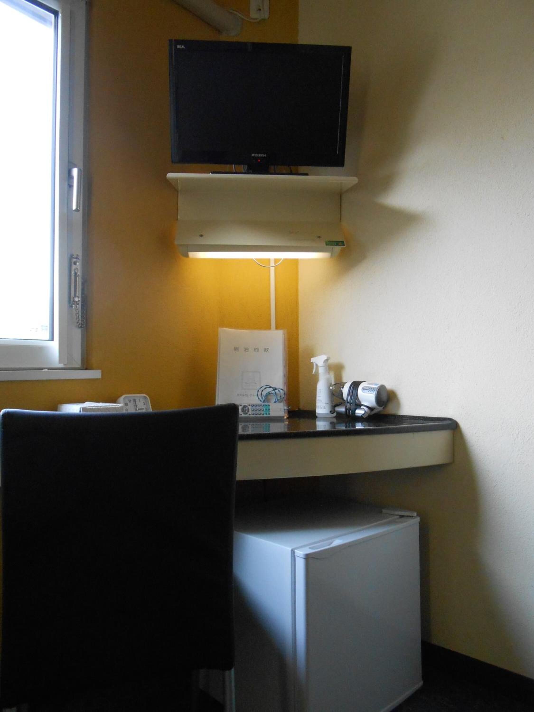 Une pièce de taille modeste comprenant un bureau, une chaise, un téléviseur fixé au-dessus du bureau, un petit réfrigérateur et une fenêtre laissant entrer la lumière naturelle.