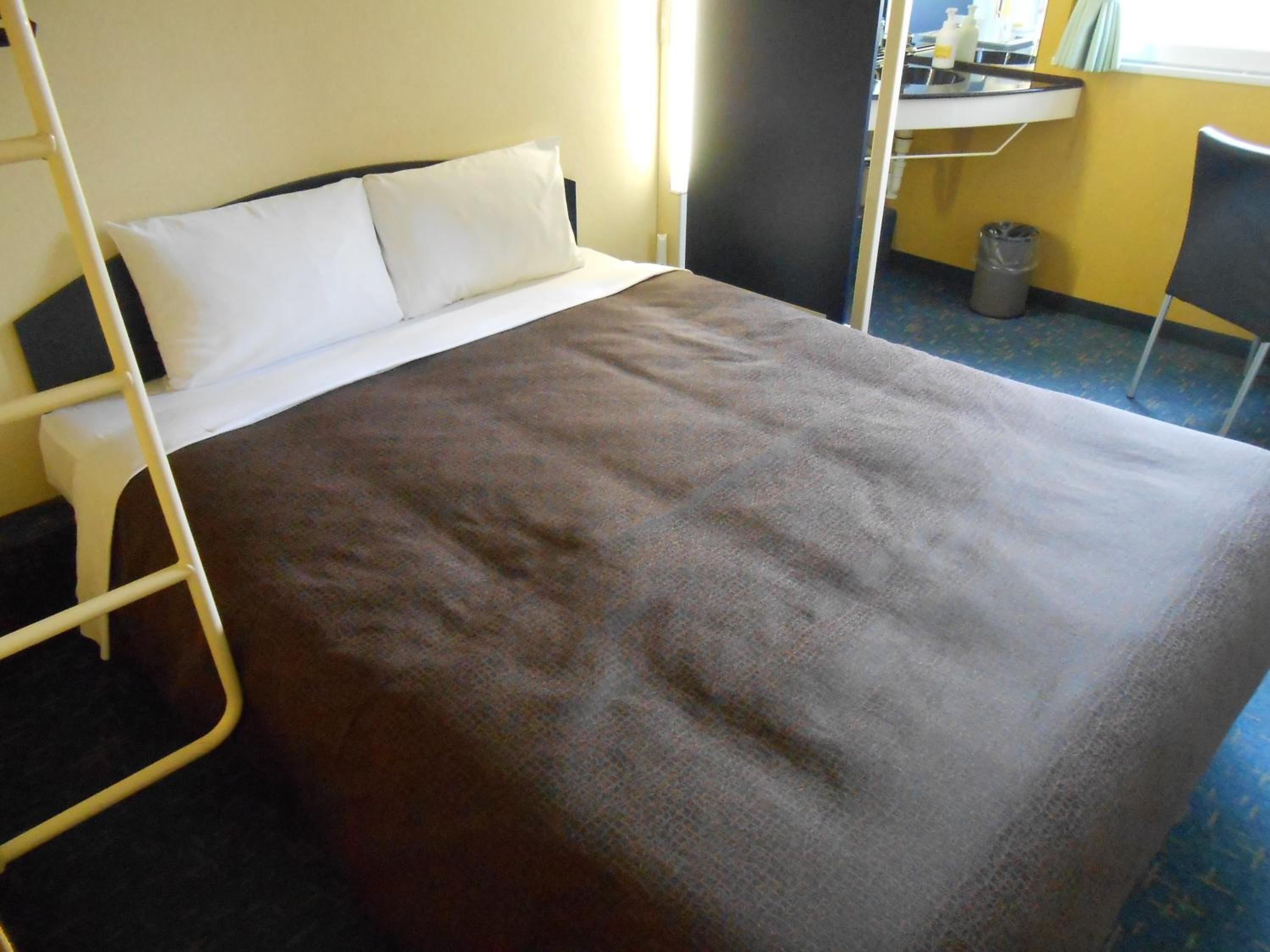 A single bed with a brown blanket, a white pillow, and a metal frame. There is a small desk with a chair and a trash can in the corner.