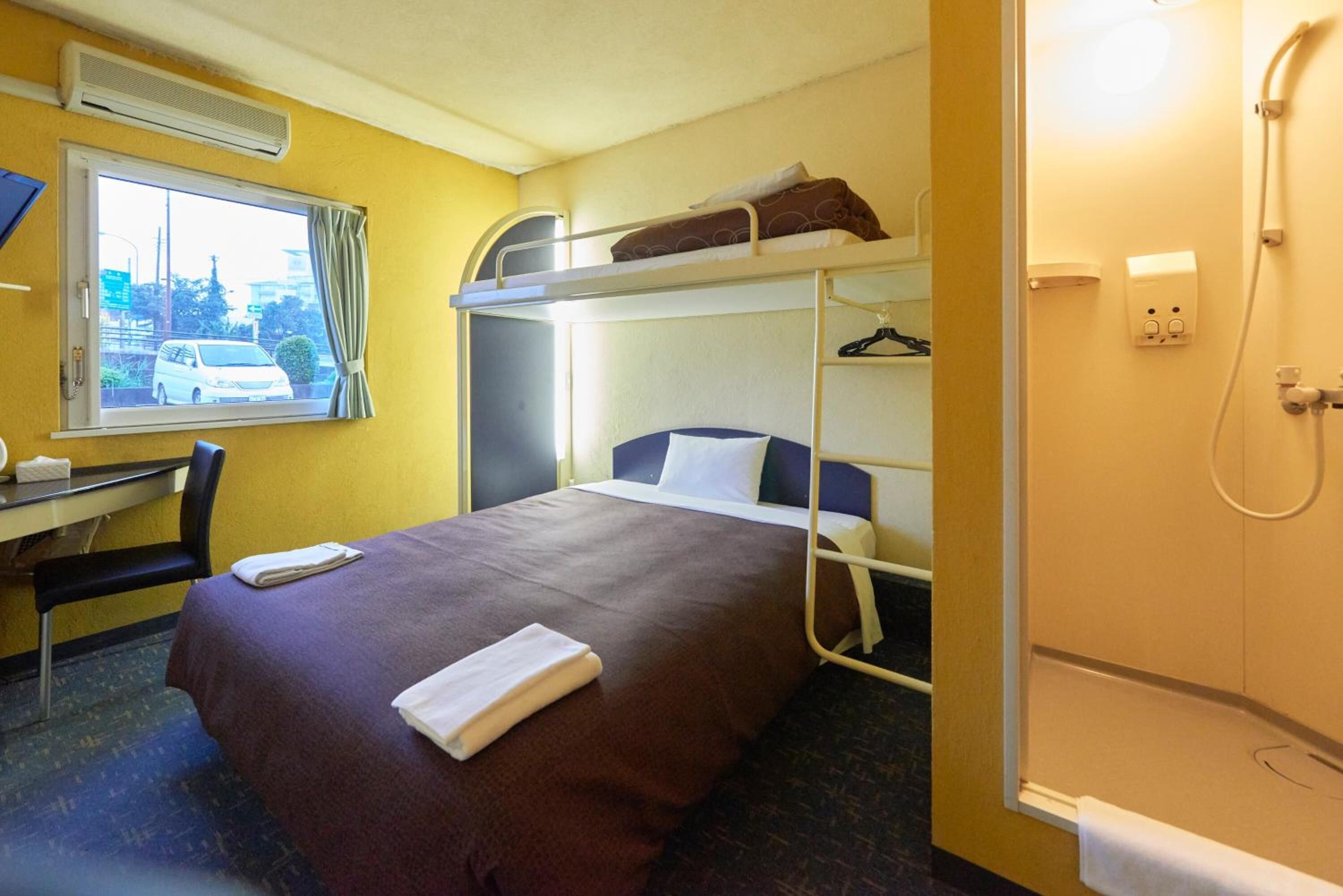 A hotel room with a bunk bed, a desk with a chair, a window with a view of a car, and a shower area.