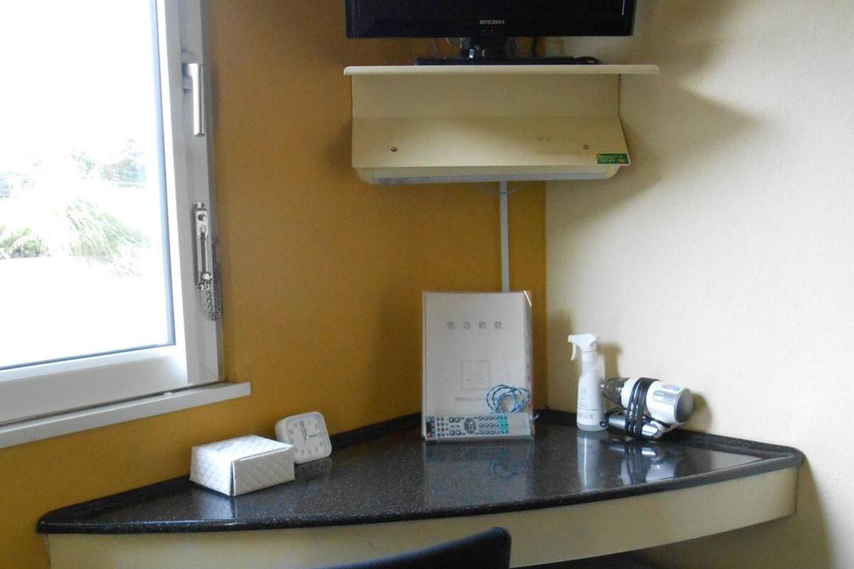 A small room with a desk, a chair, a television mounted on the wall, a small refrigerator, and a window with a view of greenery outside.