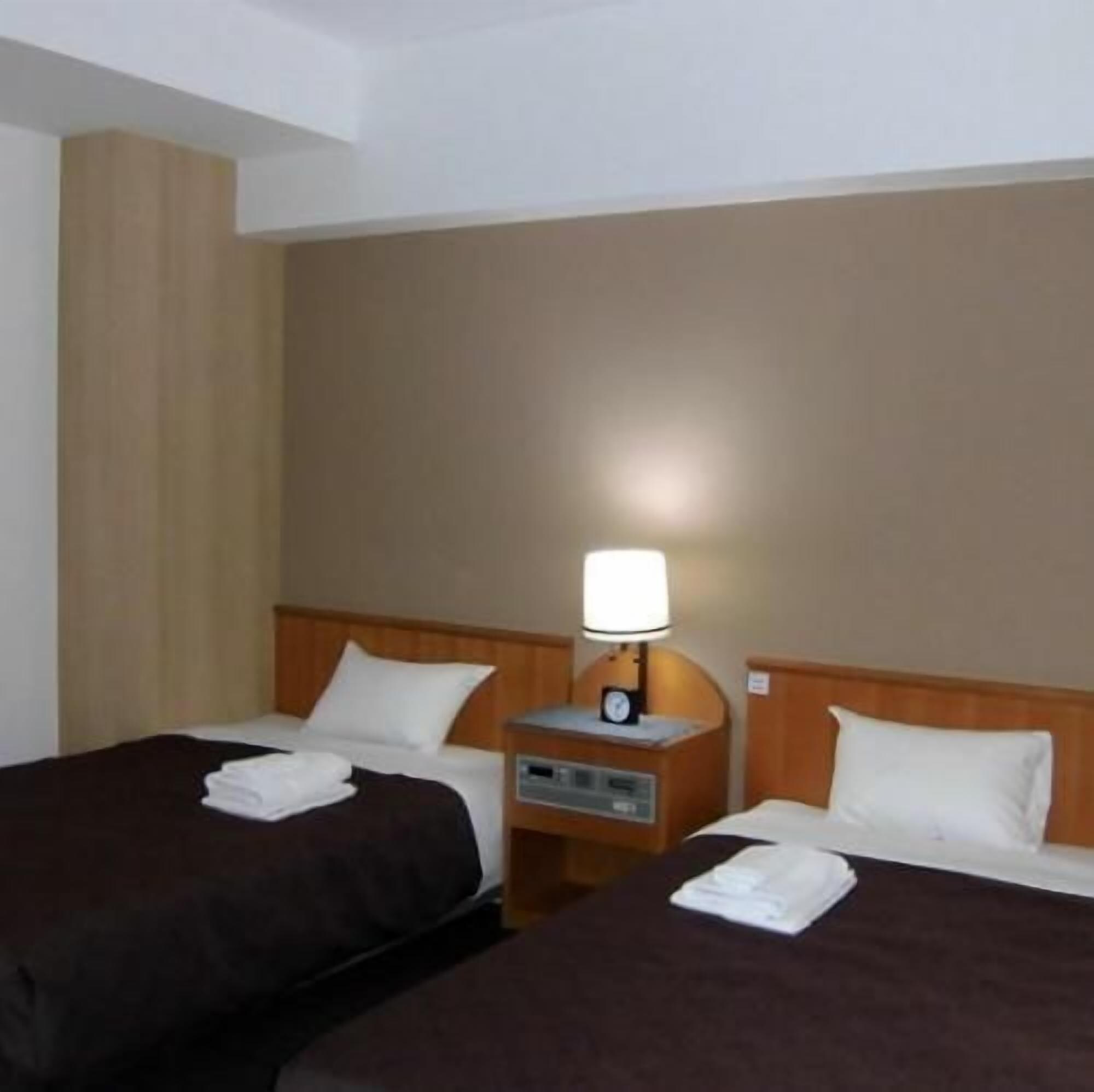 A hotel room with two single beds, a nightstand with a lamp, and a wall-mounted clock.