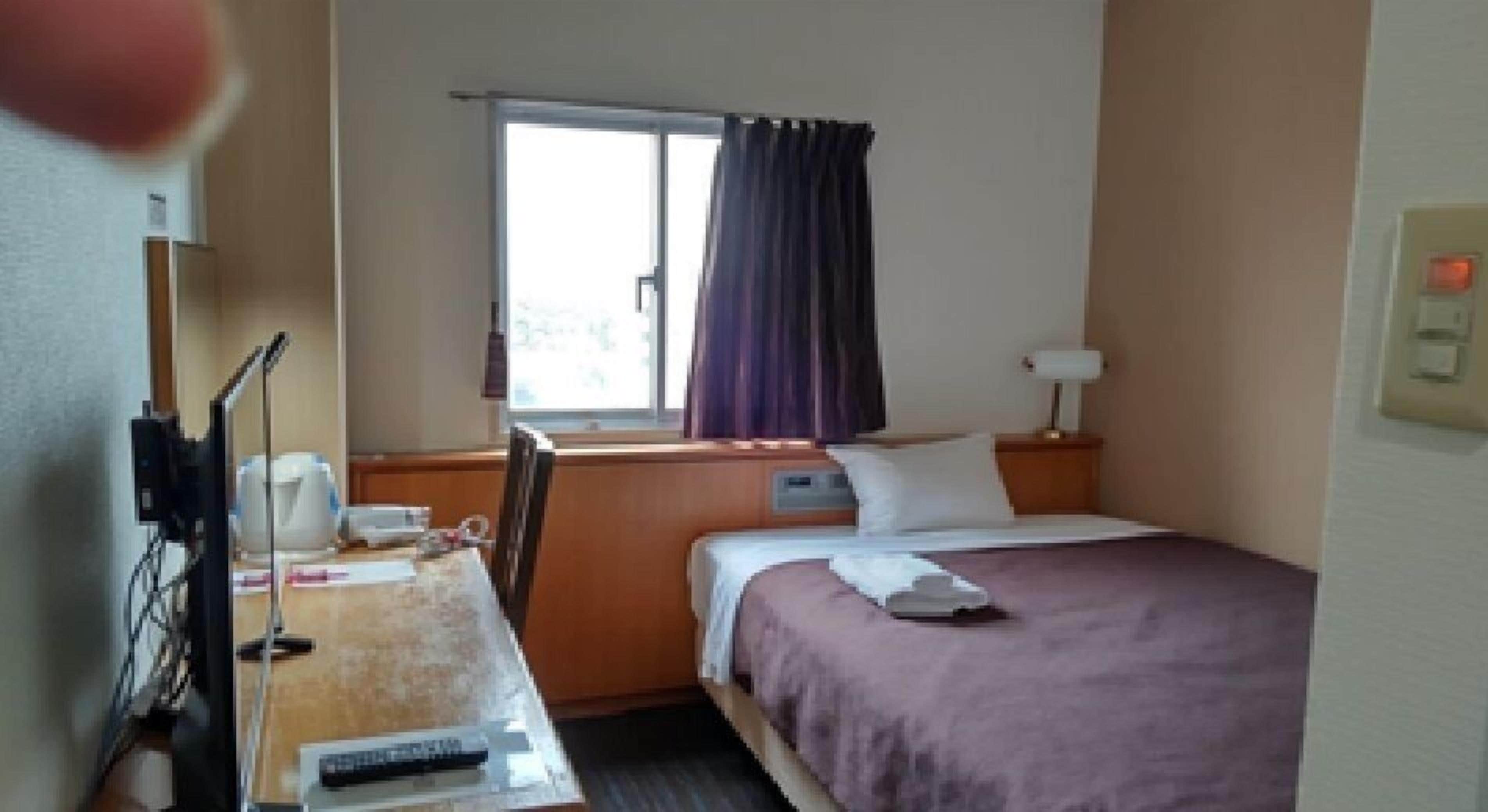 A hotel room with a bed, a desk with a computer and a chair, a window with curtains, and a nightstand with a lamp.