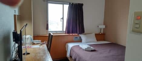 A hotel room with a bed, a desk with a computer and a chair, a window with curtains, and a nightstand with a lamp.