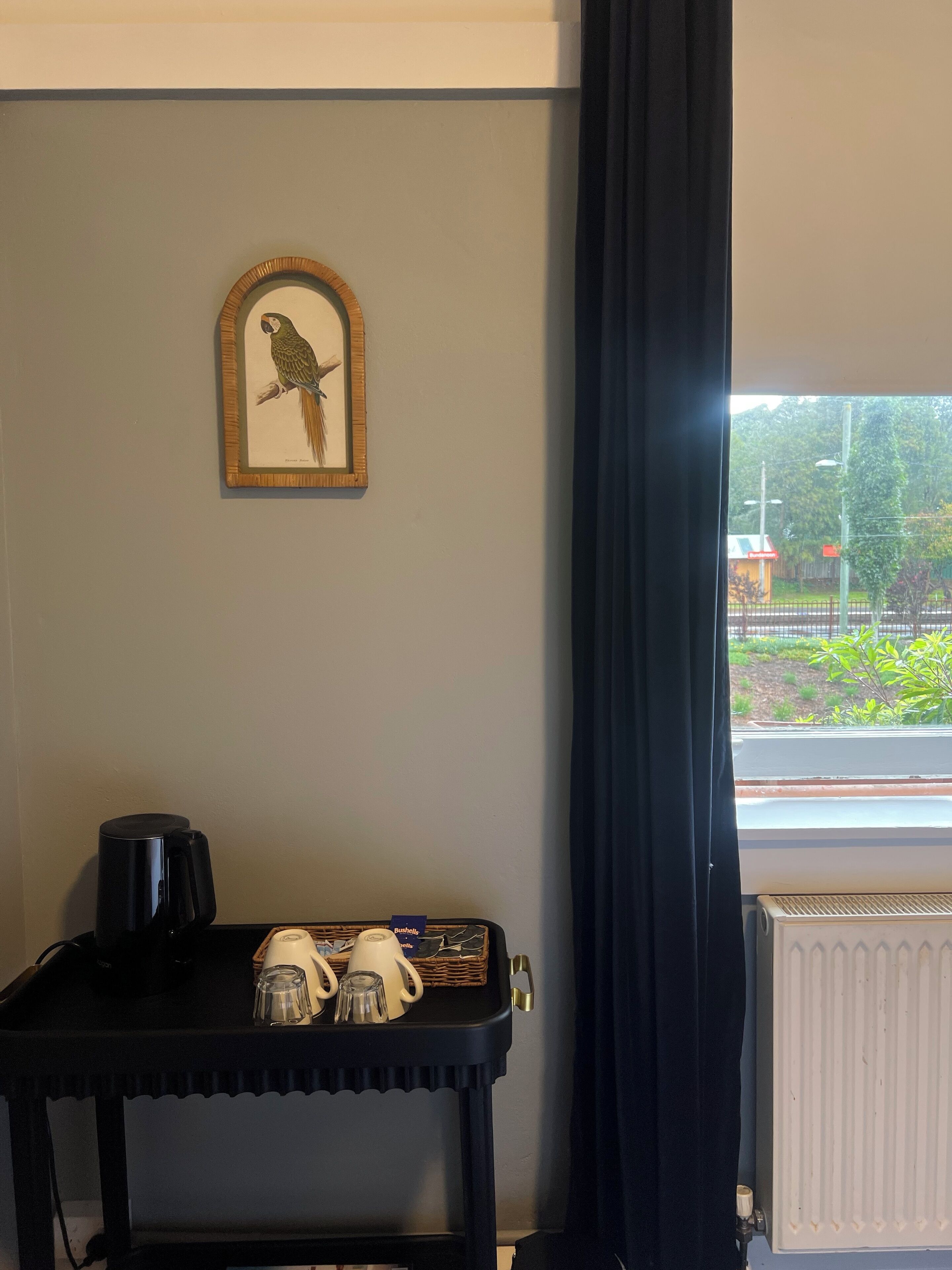 Une pièce avec une illustration d’oiseau encadrée accrochée au mur, une machine à café noire posée sur une table et une fenêtre donnant sur un paysage verdoyant et une clôture.