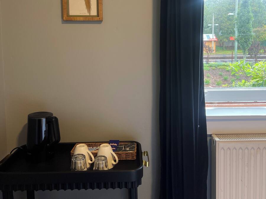 Une pièce avec une illustration d’oiseau encadrée accrochée au mur, une machine à café noire posée sur une table et une fenêtre donnant sur un paysage verdoyant et une clôture.