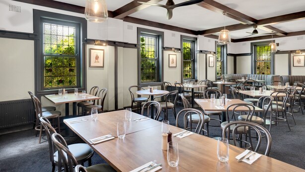 A restaurant with multiple tables and chairs, large windows with greenery visible outside, and ceiling fans. - Bundanoon Hotel (Bundanoon)