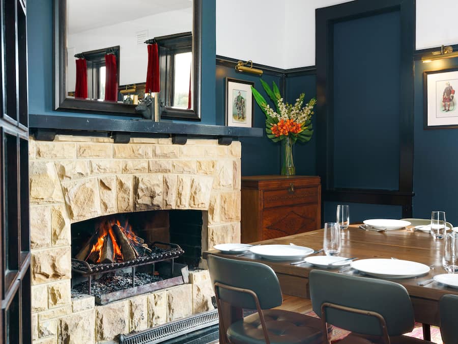 A dining area with a stone fireplace, a wooden table set for four, and a vase of flowers.