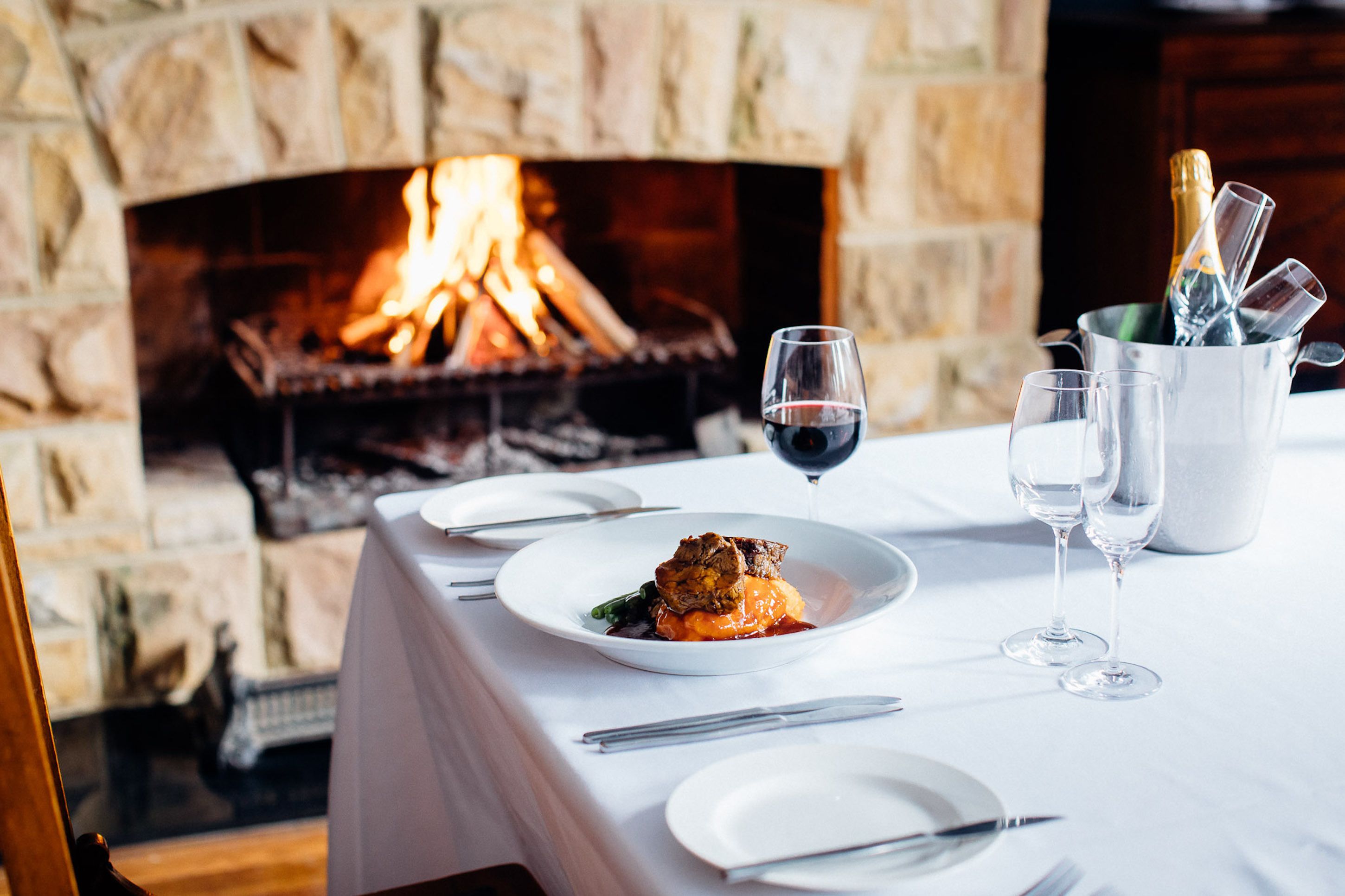 Una mesa de comedor preparada con platos, vasos y cubiertos, con una chimenea al fondo.
